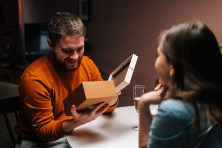 Cheerful young man opening box with received gift from loving girlfriend enjoying romantic dinner date on birthday or Valentines Day. Pretty young woman giving wrapped box with gift to husband at homeの写真素材