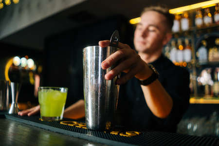 Low-angle view of professional barman making colorful alcoholic cocktail standing behind bar counter in modern dark nightclub, on blurred background of shelves with different alcoholic drinks.の写真素材