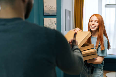 Cheerful young woman open box of hot pizza standing at entryway on apartment, smiling looking to delivery man. Back view of unrecognizable courier male delivering boxes with food to female to home.の写真素材