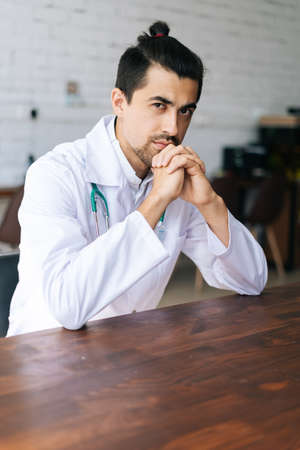 Vertical portrait of serious handsome male doctor wearing white uniform with stethoscope sitting at desk in hospital office, looking at camera. Concept of medicine and health care.の写真素材