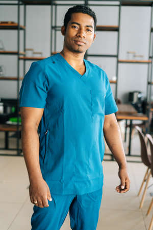 Vertical medium shot portrait of serious African American male doctor wearing blue surgeon medical uniform standing in hospital office, looking at camera. Confident practitioner posing at workplace.の写真素材