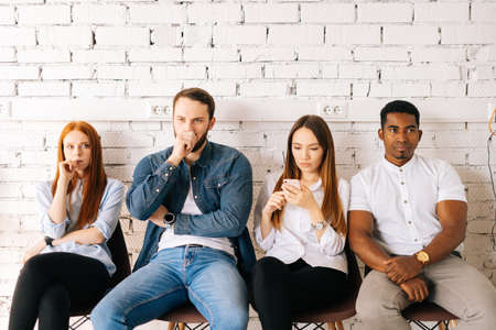 Portrait of tired young diverse multicultural job candidates in casual clothes waiting interview with hr, sitting in queue line row on chairs in modern office lobby on background of white brick wall.の写真素材