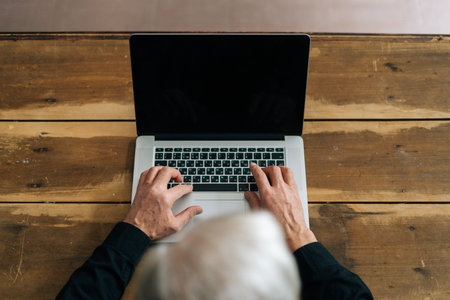 Close-up top view of unrecognizable gray-haired senior male using typing on laptop keyboard sitting at table at home office. Closeup of aged business man working on computer indoors, surfing online.の写真素材