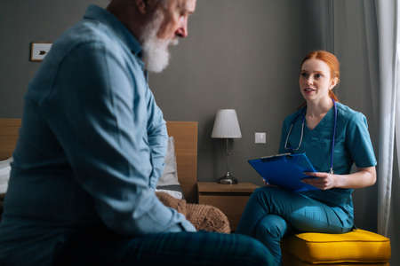 Female doctor writing down diagnosis and symptoms in medical history on clipboard, talking to sick senior male patient sitting on bed at home. Elderly man getting medical consultation in hospital.の写真素材