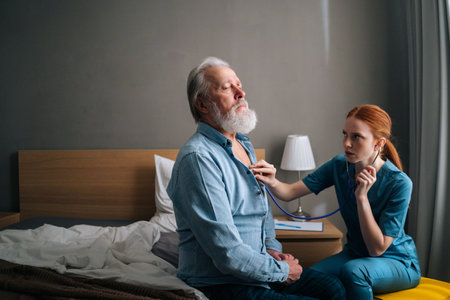 Female doctor listening with stethoscope lungs and heart of senior aged male patient sitting on bed at home. Cardiologist checking elderly man heartbeat at checkup medical visit in hospitalの写真素材