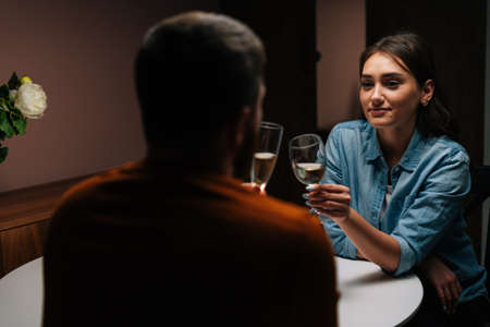 Rear view of happy young couple clinking glasses of white wine sitting at table with candles in dark cozy room. Loving man and woman celebrating anniversary, Valentines day, enjoying romantic date.の写真素材