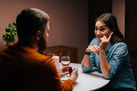 Rear view loving young man making proposal for marriage to happy pretty woman sitting at table with...