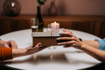 Close-up cropped shot of woman giving wrapped box with gift to man sitting at table with candles on...