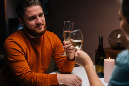 Close-up of happy young couple clinking glasses of white wine sitting at table with candles in dark cozy room. Love couple celebrating anniversary, Valentines day, enjoying romantic date.の写真素材