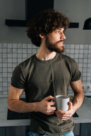 Vertical portrait of pensive young man holding in hand cup with morning coffee, thoughtful looking away, standing in kitchen with modern light interior, enjoying fresh warm drink at home office.の写真素材