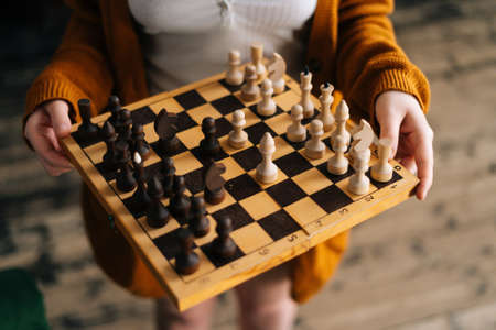 High-angle view cropped shot of unrecognizable young woman holding in hands chess board standing on vintage wooden floor in dark room, selective focus, close-up.の写真素材