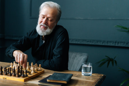 Portrait of happy bearded mature male performing move with pawn piece on wooden chessboard, selective focus. Handsome smiling gray-haired man playing chess alone at home.の写真素材