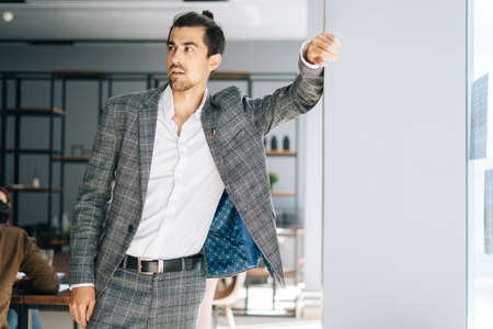 Portrait of confident elegant business man wearing fashion suit standing by window in modern office room, looking away. Front view of bearded businessman posing at workplace.の写真素材