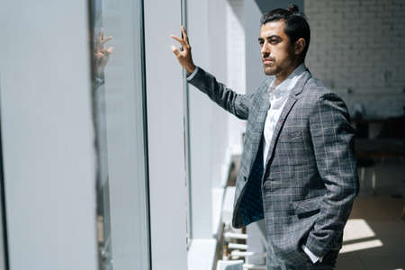 Portrait of thinking elegant business man wearing fashion suit standing in modern office room, looking out window. Front view of serious bearded businessman posing at workplace.の写真素材