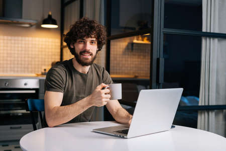 Portrait of positive bearded young business man holding in hand cup with morning coffee sitting at...