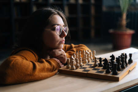 Close-up side view of pensive woman in elegant eyeglasses thinking about chess move sitting on floor in dark library room, selective focus. Pretty intelligent lady playing logical game alone at home.の写真素材