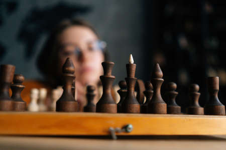 Close-up selective focus shot of unrecognizable pensive young woman in elegant eyeglasses thinking about chess move sitting on floor in dark room. Pretty intelligent lady playing logical game alone.の写真素材