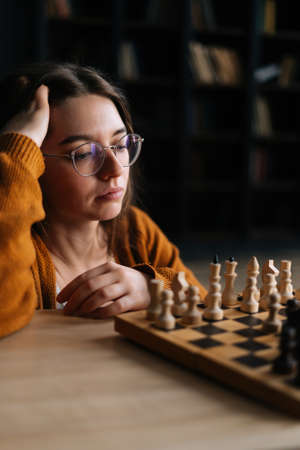 Vertical shot of thoughtful young woman in elegant eyeglasses thinking about chess move sitting in dark library room, selective focus. Pretty intelligent lady playing logical board game alone at home.の写真素材