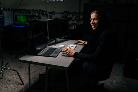 Smiling caucasian hacker man in hood breaking into corporate data servers using laptop and computers...