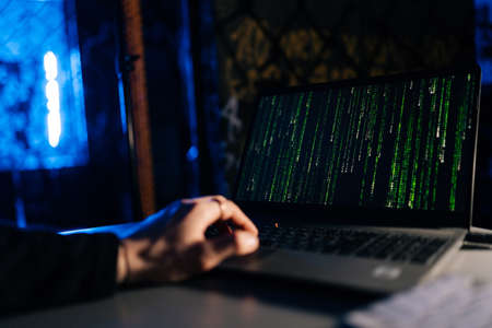 Close-up of unrecognizable hacker engaged in hacking into security system on laptop green binary code on screen with sitting at desk in dark basement room, with blue neon lights.の写真素材
