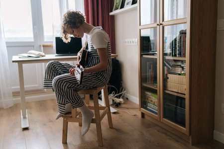 Wide shot of adorable curly little girl sitting on chair and playing on electric guitar, learning new song at home. Pretty kid teaching to play guitar in cozy bedroom with modern interior.の写真素材