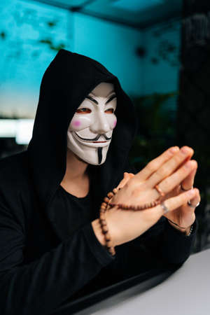 Close-up vertical shot of dangerous hacker man wearing anonymous mask and hoodie holding beads in hands and looking away sitting at table in basement room, on background of light screen of computers.の写真素材