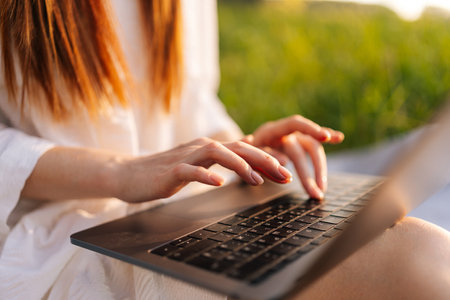 Close-up cropped shot of unrecognizable female freelancer working typing on laptop computer sitting on beautiful field with green grass, on background of warm sunlight in summer sunny day.の写真素材