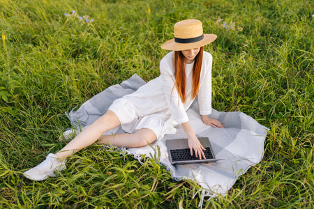 High-angle view of beautiful young woman in straw hat and white dress working on laptop computer...