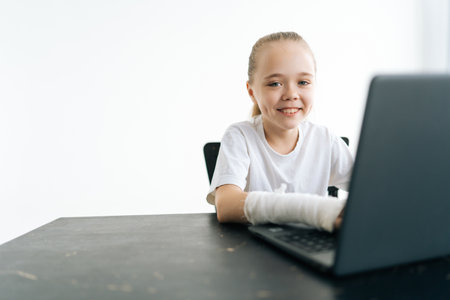 Smiling little girl with broken hand wrapped in white plaster bandage typing on laptop computer looking at camera sitting at table in light room. Concept of child insurance and healthcare.の写真素材