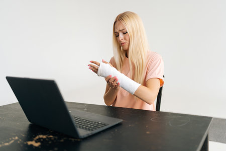 Portrait of frustrated blonde young woman with broken arm wrapped in plaster bandage having video call, online consultation using laptop computer sitting at black table, on white isolated background.の写真素材