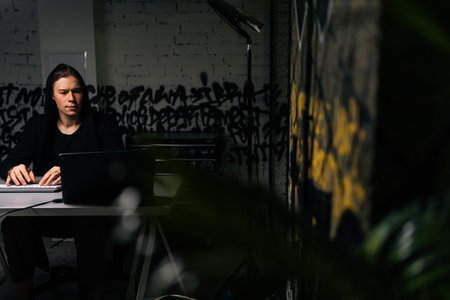 Focused hacker man in sweatshirt hacking online web site or engaging hacking into security systems, using laptop and desktop computer at desk in dark room. Concept of data thief, internet attack.の写真素材