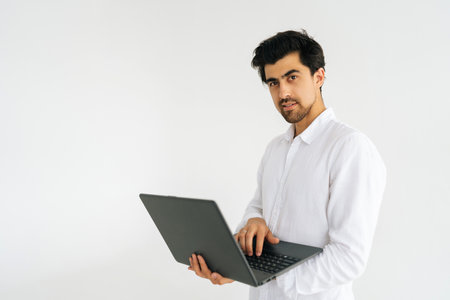 Studio portrait of handsome bearded young man posing with laptop computer and looking at camera, standing on white isolated background. Confident freelancer male holding notebook, copy space.の写真素材