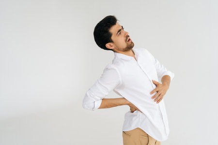 Studio portrait of unhealthy young man in shirt touching injured sore back, suffering lower lumbar...