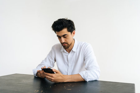 Studio portrait of serious bearded young man in shirt holding smartphone, using online mobile apps, looking on device screen, chatting in messengers sitting at table on white isolated background.の写真素材