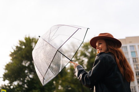 Low-angle back view of cheerful elegant young woman wearing fashion hat closing transparent umbrella...