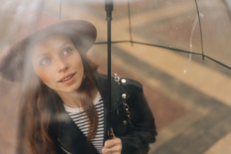 Shooting through transparent umbrella to charming redhead young woman in fashion hat standing on city street in rainy weather rain, smiling looking away. Concept of female lifestyle at autumn season.の写真素材