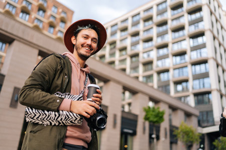 Low-angle view of cheerful bearded young man wearing stylish woman hat holding in hand camera,...