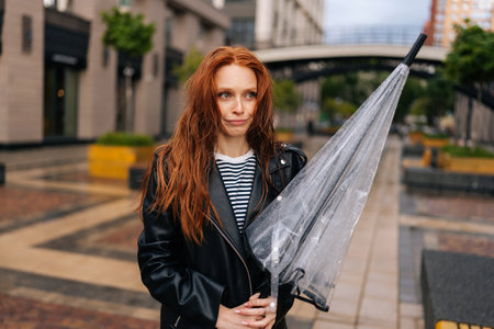 Portrait of disgruntled attractive young woman with long red-hair standing with wet, disheveled hair after being caught in cold autumn rain, holding in hand broken umbrella, sad looking away.の写真素材