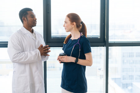 Portrait of African American black male doctor in white coat talking with positive redhead young nurse wearing blue uniform, standing on background of window at modern hospital.の写真素材