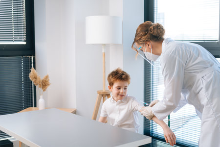 Female doctor wearing protective face mask and latex gloves vaccinating cute little child boy at hospital with modern light interior. Concept of vaccination program, prevention of infectious diseases.の写真素材