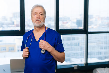 Portrait of confident senior adult male doctor in medical uniform standing in hospital office on...