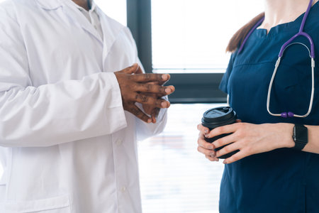 Close-up cropped shot of unrecognizable African-American male doctor in white coat talking with young female nurse in blue uniform holding coffee cup, standing on background of window at hospital.の写真素材