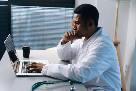 Side view of confused black male doctor wearing white coat uniform thinking looking at laptop computer screen sitting at desk in light medic clinic office room, on background of window.の写真素材