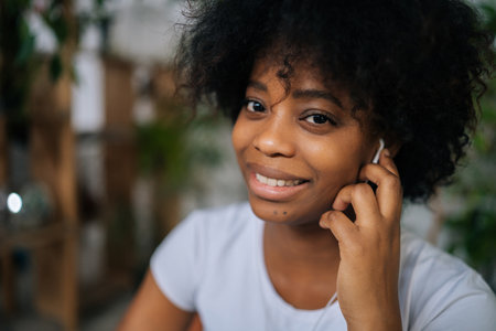 Close-up side view of smiling African-American young woman enjoying quality sound, favourite music through earphones. Happy black female listen podcast or classic tune using modern in-ear device.の写真素材