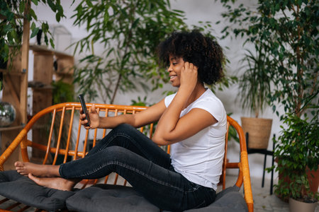 Portrait of happy young African American woman listening music from mobile phone, choosing next song looking to screen sitting on couch at home in room with modern green biophilic interior design.の写真素材