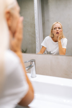 Vertical portrait of sad young woman with blonde hair touching face and looking at reflection in mirror with worried face in bathroom with modern light interior. Idea of body and skin careの写真素材