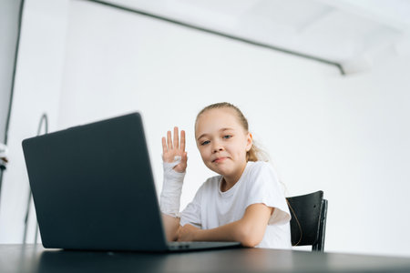 Positive little girl with broken hand wrapped in white plaster bandage showing hello gesture looking at camera sitting at desk with laptop in light room. Concept of child insurance and healthcare.の写真素材