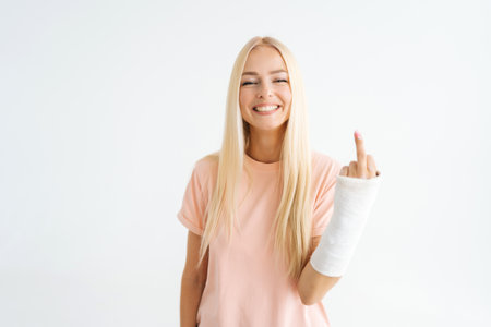 Cheerful blonde young woman showing middle finger, rude gesture with broken arm wrapped in plaster bandage standing on white isolated background, looking at camera. Concept of insurance and healthcareの写真素材