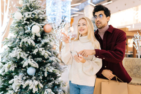 Portrait of smiling young couple using smartphone together standing with paper bags in hall of celebrate shopping mall in Christmas eve, on background of bright beautiful xmas decorations.の写真素材