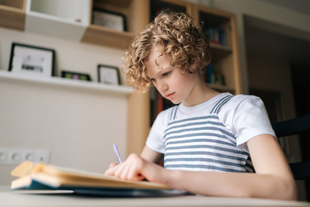 Low-angle view of concentrated little child schoolgirl writing in exercise book doing some homework, sitting at desk near window. Cute curly small school girl studying alone making notes at home.の写真素材